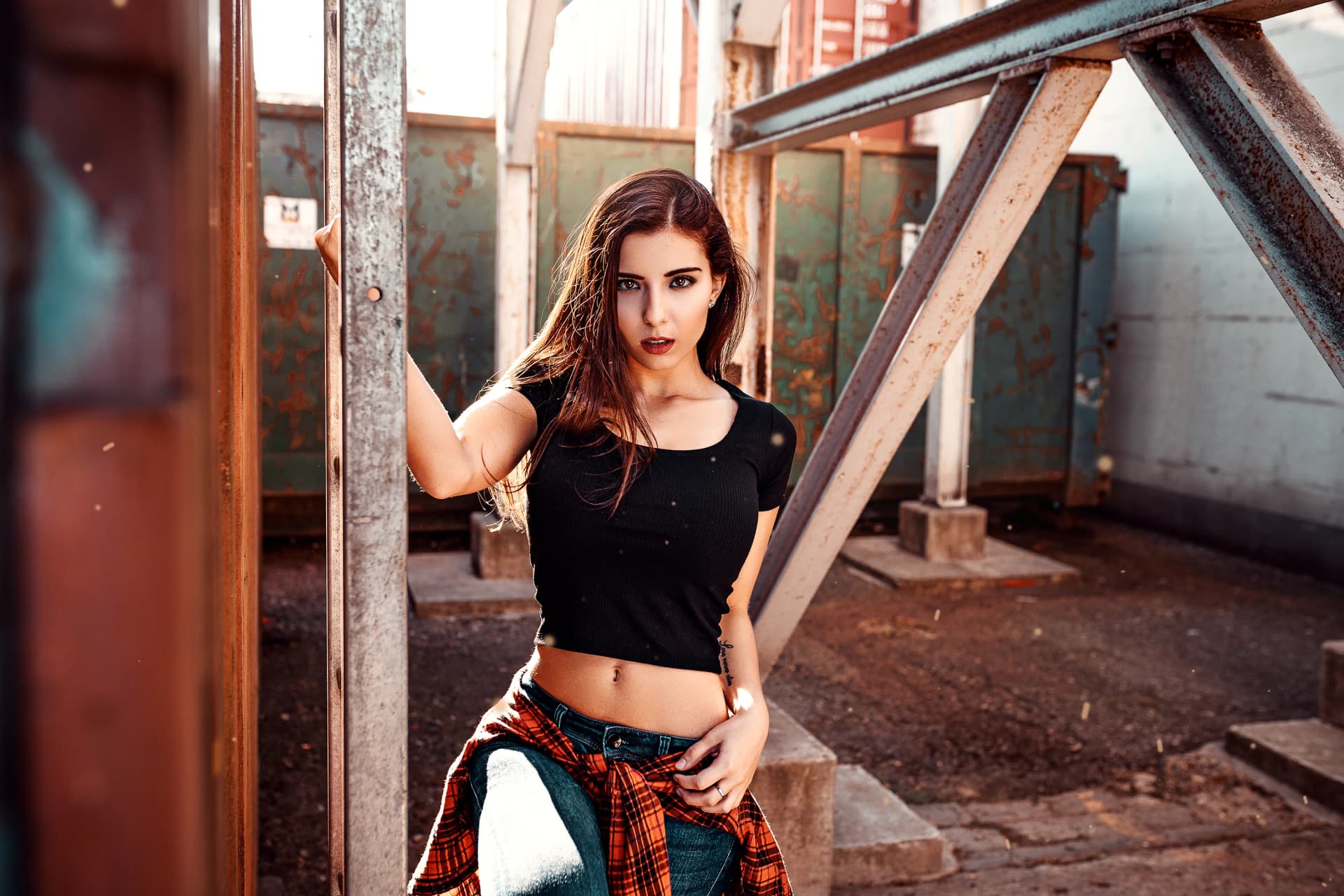 A portrait of a dark-haired young woman, dressed in summer fashion, in an urban industrial location with containers. The particles in the air and the bright sunlight give the image dynamism and a modern, contrasting atmosphere with a pinch of sexiness.